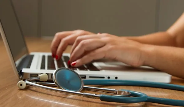 Hands typing on a laptop beside a stethoscope on a desk